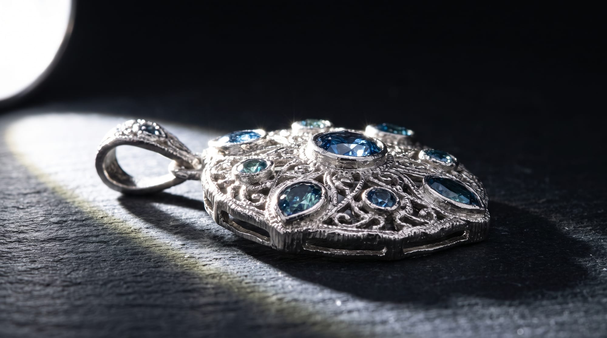 A round white-metal pendant of openwork filigree set with blue sapphires of several cuts around a larger oval centre stone, on slate.